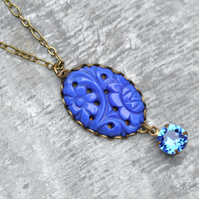 K&ouml;nnte beinhalten: Eine blaue Blumen-Halskette mit einem blauen Kristall-Anh&auml;nger. Der Anh&auml;nger ist aus geschnitztem Glas und hat eine Messingkette.