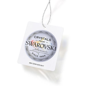 K&ouml;nnte beinhalten: Ein wei&szlig;es Etikett mit einem silbernen Kreis, der den Text "Crystals from Swarovski" und "Since 1895" enth&auml;lt. Der Text "ABW1 ZZWW WZT0 OK1T" ist unter dem Kreis gedruckt.
