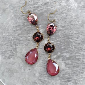 May include: A pair of drop earrings featuring three faceted crystals in shades of pink and deep red, set in antique brass-toned metal settings. Each earring hangs from a simple hook wire, showcasing a teardrop-shaped top, a square middle, and a larger teardrop-shaped bottom crystal.