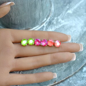 May include: Four pairs of small round stud earrings in a rainbow gradient of colors. The earrings are green, pink, red, and orange. The earrings are on a hand.