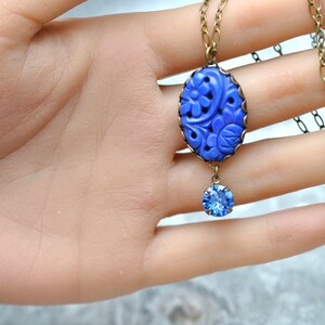 K&ouml;nnte beinhalten: Eine blaue, geschnitzte Anh&auml;nger-Kette mit einem blauen Kristall-Tropfen. Der Anh&auml;nger ist oval geformt und hat ein florales Design. Die Kette ist aus einer Messingkette gefertigt.