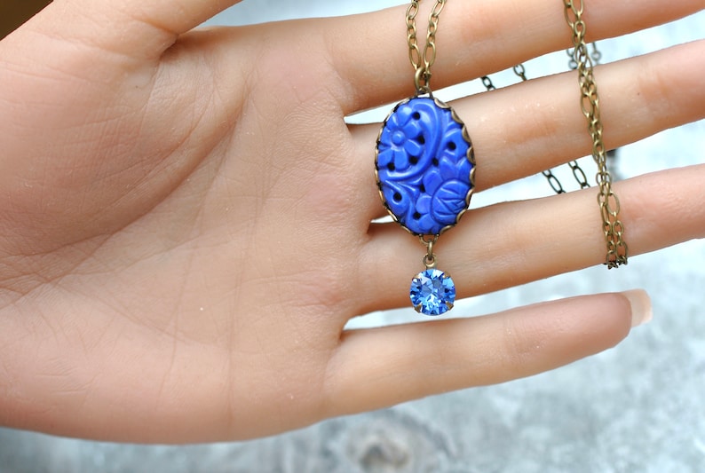K&ouml;nnte beinhalten: Eine blaue, geschnitzte Anh&auml;nger-Kette mit einem blauen Kristall-Tropfen. Der Anh&auml;nger ist oval geformt und hat ein florales Design. Die Kette ist aus einer Messingkette gefertigt.