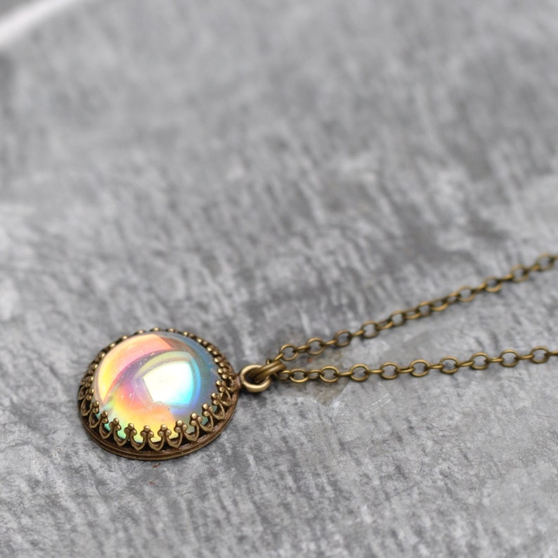 May include: A round pendant necklace with a shimmering, iridescent center. The pendant is set in a bronze-colored, ornate frame and hangs from a delicate chain. The necklace is displayed on a gray, textured surface.