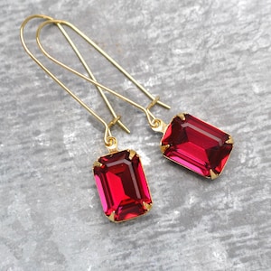 May include: A pair of gold-tone earrings featuring large, rectangular, ruby-red gemstones. The gemstones are set in gold-colored frames and suspended from long, curved gold-tone ear wires. The earrings are displayed against a gray background.