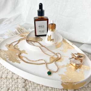 May include: White oval tray with gold leaf accents, featuring a gold chain necklace with a green stone pendant, gold hoop earrings, a small white ceramic ring holder with a brown top, and a brown glass bottle with a dropper labelled "Argan Oil Made in Morocco PEAU".