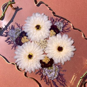 K&ouml;nnte beinhalten: Ein rosa Harztablett mit goldfarbenem Rand und einem goldfarbenen Griff. Das Tablett ist mit wei&szlig;en Blumen und violetten Trockenblumen gef&uuml;llt.