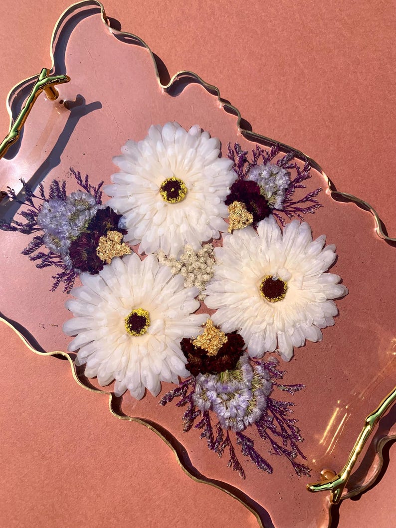 K&ouml;nnte beinhalten: Ein rosa Harztablett mit goldfarbenem Rand und einem goldfarbenen Griff. Das Tablett ist mit wei&szlig;en Blumen und violetten Trockenblumen gef&uuml;llt.