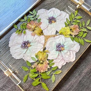 K&ouml;nnte beinhalten: Ein klares Acryltablett mit goldener Einfassung, verziert mit gepressten Blumen in Rosa-, Lila-, Gelb- und Gr&uuml;nt&ouml;nen. Die Blumen sind kreisf&ouml;rmig angeordnet und bilden ein wundersch&ouml;nes und zartes Design.