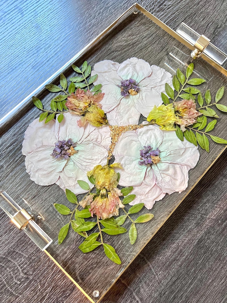 K&ouml;nnte beinhalten: Ein klares Acryltablett mit goldener Einfassung, verziert mit gepressten Blumen in Rosa-, Lila-, Gelb- und Gr&uuml;nt&ouml;nen. Die Blumen sind kreisf&ouml;rmig angeordnet und bilden ein wundersch&ouml;nes und zartes Design.