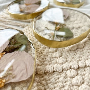 K&ouml;nnte beinhalten: Vier runde, transparente Harz-Untersetzer mit goldenen Metallr&auml;ndern. Jeder Untersetzer zeigt gepresste getrocknete Blumen und Gr&uuml;npflanzen.