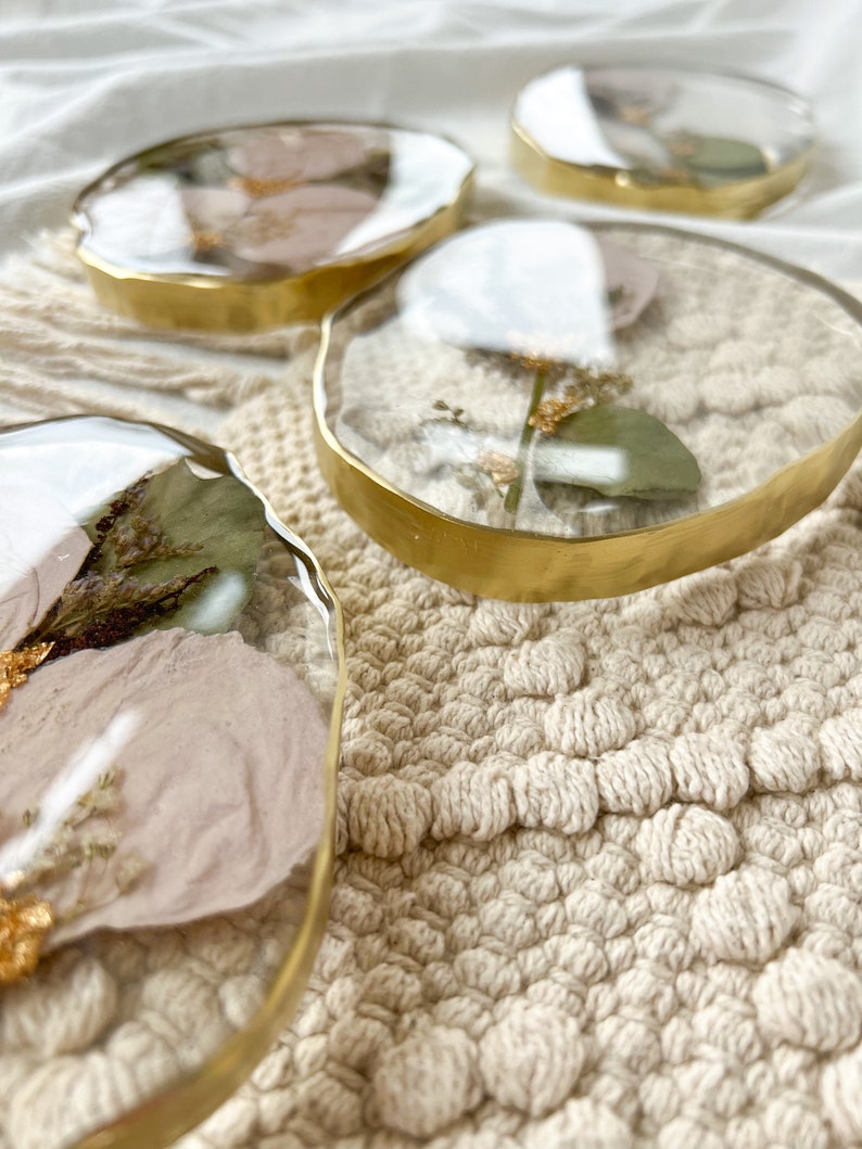 K&ouml;nnte beinhalten: Vier runde, transparente Harz-Untersetzer mit goldenen Metallr&auml;ndern. Jeder Untersetzer zeigt gepresste getrocknete Blumen und Gr&uuml;npflanzen.