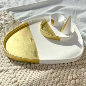 May include: A white and gold ceramic tray with two smaller white ceramic dishes. The tray is oval shaped and has a gold rim. The smaller dishes are a round bowl and a cone shaped dish.