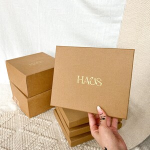 K&ouml;nnte beinhalten: Stapel aus vier braunen Pappkartons mit dem Wort "HAUS" in Gold auf dem oberen Karton.