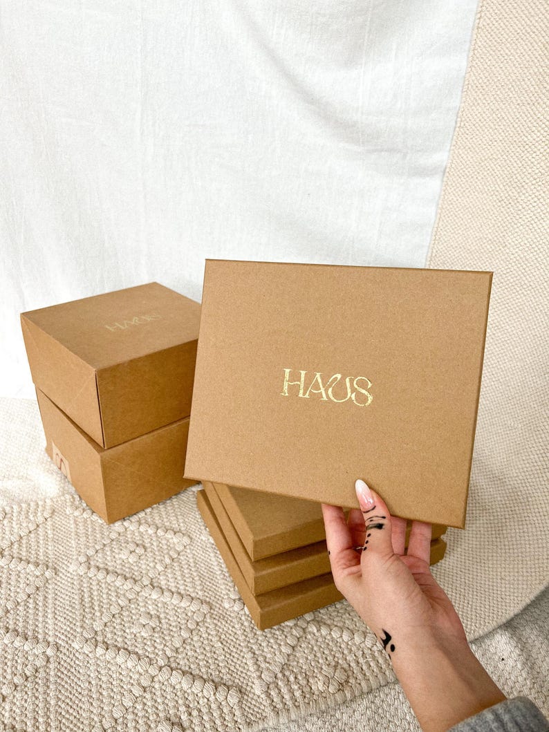 K&ouml;nnte beinhalten: Stapel aus vier braunen Pappkartons mit dem Wort "HAUS" in Gold auf dem oberen Karton.