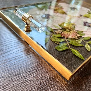 K&ouml;nnte beinhalten: Ein goldfarbener Tablett mit einer durchsichtigen Glasplatte, verziert mit gepressten Blumen und Gr&uuml;npflanzen. Das Tablett hat einen durchsichtigen Acryl-Griff mit einem goldenen Akzent.