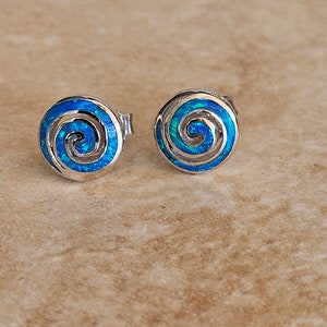 Könnte beinhalten: Ein Paar silberne Ohrstecker mit einem Spiraldesign. Die Ohrringe haben einen leuchtenden blau-grünen Opal-ähnlichen Einsatz in einem kreisförmigen Rahmen. Die Ohrstecker sind vor einem neutralen Hintergrund platziert.