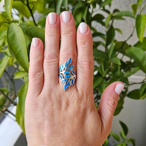 Könnte beinhalten: Ein silberner Ring mit Blattdesign, besetzt mit blauen Opalsteinen. Der Ring wird auf einer Hand mit manikürten Nägeln vor einem Hintergrund aus grünem Laub gezeigt.
