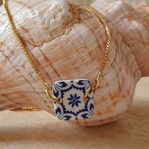 Könnte beinhalten: Eine goldene Kettenkette mit einem blauen und weißen Keramik-Anhänger mit floralem Design. Der Anhänger ist quadratisch und hat einen goldenen Rahmen.