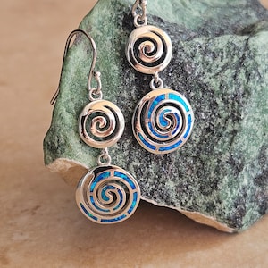 Könnte beinhalten: Silberfarbene Ohrhänger mit einem Spiraldesign. Jeder Ohrring hat drei kreisförmige Elemente, wobei die unteren zwei Kreise einen blauen, opalartigen Einsatz enthalten. Die Ohrringe werden vor einem grünen und beigen Hintergrund präsentiert.