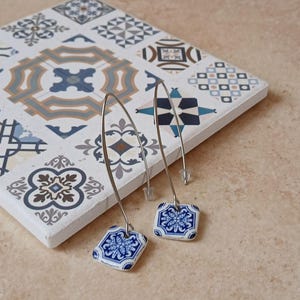Könnte beinhalten: Ein Paar Hängeohrringe mit blauen und weißen Keramik-Anhängern, die von Fliesen inspiriert sind. Die Anhänger zeigen ein geometrisches Muster mit einem floralen Motiv. Die Ohrringe hängen an silberfarbenen Haken.