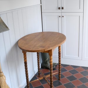 Puede incluir: Mesa auxiliar ovalada de madera con patas torneadas. La mesa tiene un acabado marrón claro y está colocada en una esquina contra una pared blanca. Las patas están talladas. La mesa mide unos 76 cm de altura.