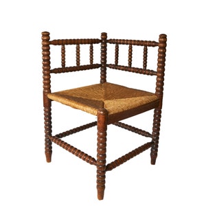 Puede incluir: Una silla de esquina construida en madera de color marrón oscuro con un diseño de husos torneados que presenta segmentos redondeados. El asiento está elaborado con fibra natural tejida en tono beige, formando un patrón cuadrado.