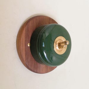 Op de afbeelding: Een ronde wandschakelaar met een donkergroene glanzende keramische basis gemonteerd op een ronde houten achterplaat. Het midden van de schakelaar heeft een goudkleurige metalen tuimelschakelaar omringd door een bijpassende ronde metalen plaat.