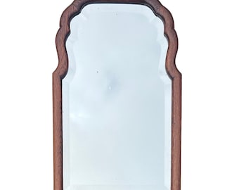May include: A rectangular mirror with a decorative, scalloped top edge. The frame is made of dark brown wood. The mirror's shape is reminiscent of an arched window. The glass is clear and reflects light.