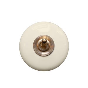 Op de afbeelding: Een ronde, gebroken witte keramische kastknop met een gladde, glanzende afwerking. Het midden is voorzien van een cirkelvormige metalen basisplaat in een gedempte messingkleur, rondom een kleine, uitstekende metalen pin in het midden.