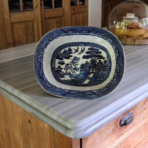 Op de afbeelding: Een ovale keramieken serveerschaal met een klassiek blauw-wit wilgenpatroon. Het ontwerp toont een gedetailleerd landschap met pagodes, wilgen, vogels en een brug. De rand is versierd met ingewikkelde geometrische en florale motieven.