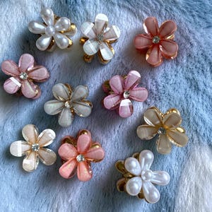 May include: A selection of flower-shaped hair clips in shades of pink, white, and gold. Some clips have pearl-like beads, others a clear, sparkling centre. The clips are displayed on a soft, blue and white textured surface.