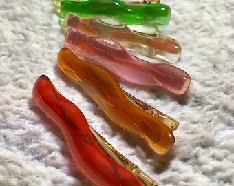 May include: A set of six colorful hair clips. The clips are made of clear plastic and have a wavy, twisted design. The colors are yellow, green, pink, orange, and red.
