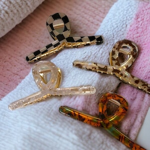 Può includere: Quattro diverse mollette per capelli in vari motivi. Le mollette sono in plastica e hanno una finitura dorata. I motivi includono un motivo a scacchi, un motivo trasparente, un motivo marrone e bianco e un motivo marrone e arancione.