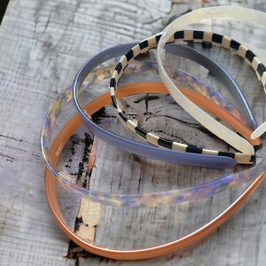 May include: Four plastic headbands in different colours and patterns. The headbands are stacked on top of each other. The headband on the bottom is orange, the next is clear with a floral pattern, the next is blue with a black and white chequered pattern, and the top headband is clear.