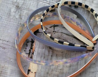 Puede incluir: Cuatro diademas de plástico en diferentes colores y patrones. Las diademas están apiladas una encima de la otra. La diadema de abajo es naranja, la siguiente es transparente con un patrón floral, la siguiente es azul con un patrón a cuadros blanco y negro, y la diadema de arriba es transparente.