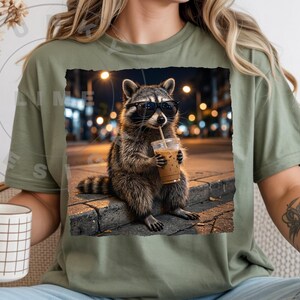May include: Olive green t-shirt featuring a graphic of a raccoon wearing sunglasses and drinking iced coffee. The raccoon is sitting on a curb at night with city lights in the background. The shirt has a relaxed fit.