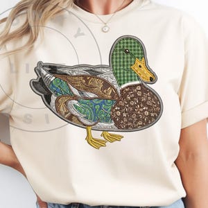 Peut inclure: T-shirt crème avec un motif de canard brodé. Le canard a une tête à carreaux verts, un bec jaune et un corps composé de tissus à motifs bruns, bleus et verts. Le motif est centré sur le t-shirt, qui a des manches retroussées.