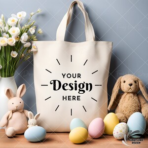 May include: A white canvas tote bag with black text that says "YOUR Design HERE" surrounded by a circle of dashes. The bag is sitting on a wooden table with Easter eggs, a bouquet of white flowers, and two plush bunny rabbits.