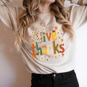 Puede incluir: Sudadera beige con la frase bordada "Give Thanks" en colores vibrantes. El diseño incluye motivos florales y hojas, una "G" con estampado de leopardo y un lazo. La sudadera es de manga larga y cuello redondo.