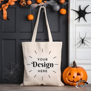 K&ouml;nnte beinhalten: Eine cremefarbene Tragetasche mit dem Text "YOUR Design HERE" in Schwarz, die an einer schwarzen T&uuml;r h&auml;ngt. Halloween-Dekorationen umfassen orangefarbene K&uuml;rbisse, Spinnen und einen geschnitzten K&uuml;rbis.
