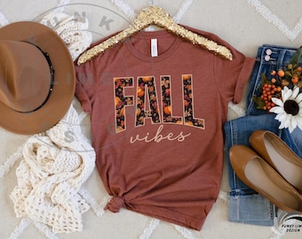 May include: A terracotta-coloured t-shirt with the words "FALL vibes" in a seasonal design. The letters are filled with a pattern of pumpkins and leaves. The shirt is paired with a brown hat, jeans, and flats.