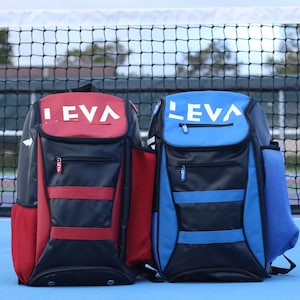 May include: Two LEVA backpacks, one red and black, the other blue and black, are displayed on a blue surface. The backpacks have the word "LEVA" in white on the top. Each backpack has a side pocket and a front zipper pocket.
