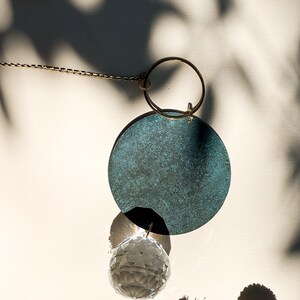 Puede incluir: Adorno colgante decorativo con un disco grande y texturizado de color verde azulado, una bola de cristal transparente y un anillo dorado. El adorno cuelga de una delicada cadena dorada. Flores secas en primer plano.