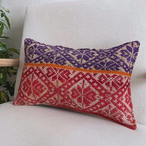 Op de afbeelding: Een rechthoekig sierkussen met een geweven textielpatroon. Het bovenste gedeelte toont een geometrisch ontwerp in paars en beige, gescheiden door een dunne oranje horizontale streep van het onderste gedeelte met een rood en beige patroon.