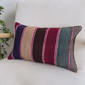 Op de afbeelding: Een rechthoekig sierkussen van geweven stof met verticale strepen in tinten dieppaars, beige, donkerbruin, magenta en gedempt groen. De randen zijn afgewerkt met een fijn, donkerbruin en wit gestreept patroon aan de zijkanten.