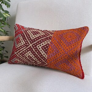Op de afbeelding: Een rechthoekig sierkussen met een geweven textielhoes in een gespleten ontwerp. De linkerkant toont een geometrisch ruitpatroon in donkerrood en crème, terwijl de rechterkant een vergelijkbaar patroon in levendig oranje en paars bevat.
