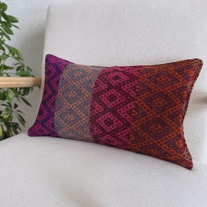 Op de afbeelding: Een rechthoekig sierkussen met een geweven geometrisch ruitpatroon in een levendig kleurenpalet van paars, magenta, grijs en gebrand oranje. Het textiel vertoont een ontwerp met meerdere panelen en verschillende verticale kleursecties.