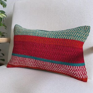 Op de afbeelding: Een rechthoekig sierkussen met een geweven textuur en horizontale strepen in de kleuren groenblauw, rood, oranje en crème. Het ontwerp bestaat uit afwisselende effen kleurstroken en gedetailleerde gespikkelde patronen over het gehele oppervlak.