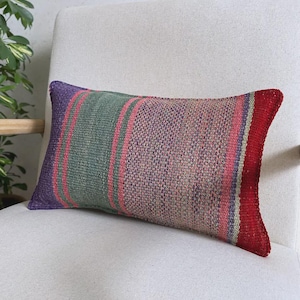 Op de afbeelding: Een rechthoekig sierkussen met een geweven textielpatroon met verticale strepen in tinten paars, saliegroen, roze en een effen rode rand aan de rechterkant. De stof heeft overal een getextureerd, meerkleurig uiterlijk.