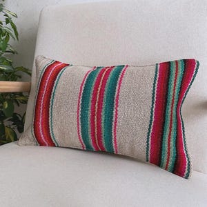 Op de afbeelding: Een rechthoekig sierkussen met een geweven beige basis en verticale strepen in levendige tinten magenta, groenblauw en oranje. De stof heeft een zichtbaar geweven textuur en het kussen is afgewerkt met strakke, rechte randen aan alle kanten.